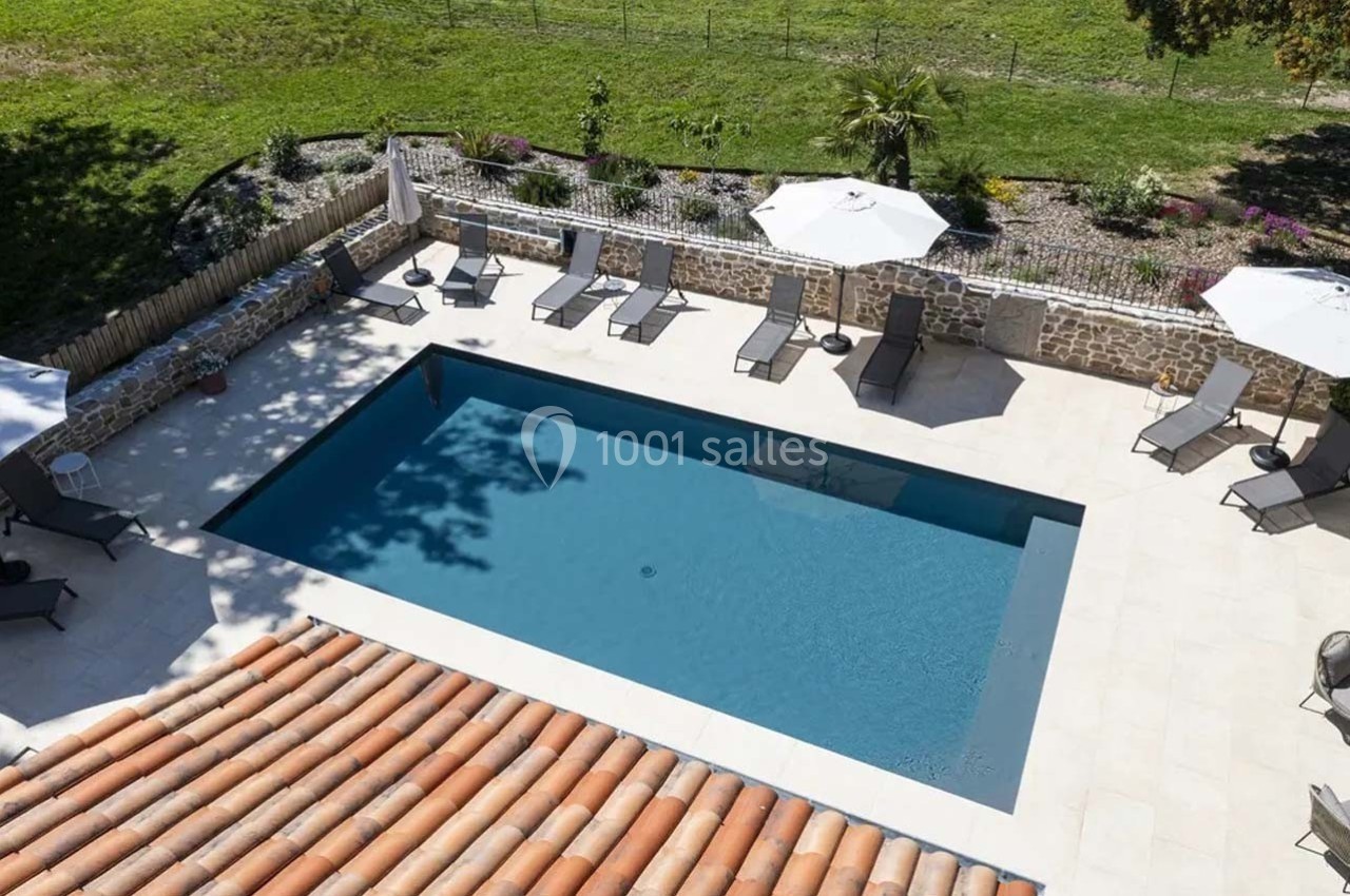Vue aérienne d'une piscine rectangulaire entourée de transats, parasols et d'un jardin clos par un muret en pierre.