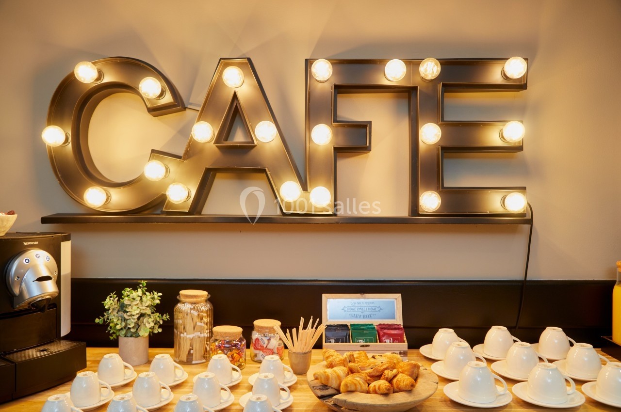 Buffet avec machine à café, tasses, viennoiseries, thés et décoration lumineuse formant le mot ’Café’.