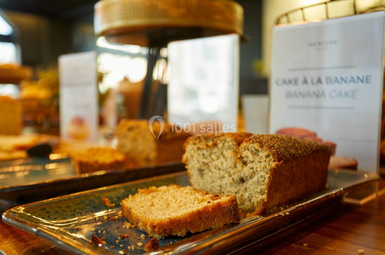 Tranche de cake à la banane posée sur un plat en céramique, avec d'autres desserts en arrière-plan flou.