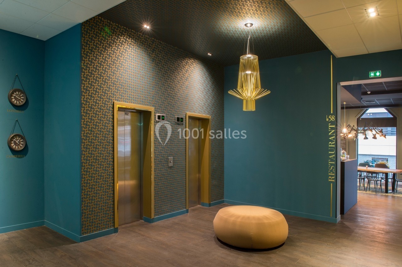 Entrée moderne avec deux ascenseurs dorés, un pouf beige, et un accès vers un restaurant indiqué sur le mur.