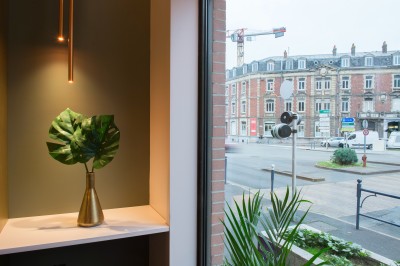 Miniature Location salle Arras (Pas-de-Calais) - Mercure Arras Centre Gare #15 Feuilles vertes dans un vase doré sur une étagère près d'une fenêtre donnant sur une rue urbaine avec bâtiments et grue.