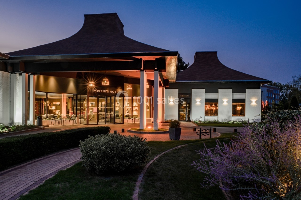 Location salle Marcq-en-Baroeul (Nord) - Mercure Lille Marcq en Baroeul #2 Entrée d'un hôtel moderne éclairée en soirée, avec jardin aménagé et chemin pavé au premier plan.