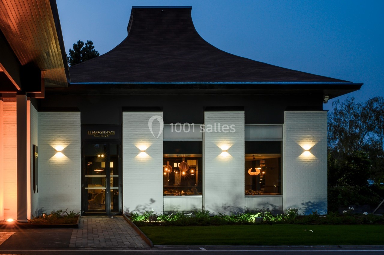 Façade d'un restaurant avec murs blancs, fenêtres éclairées et toit sombre, photographiée à la tombée de la nuit.