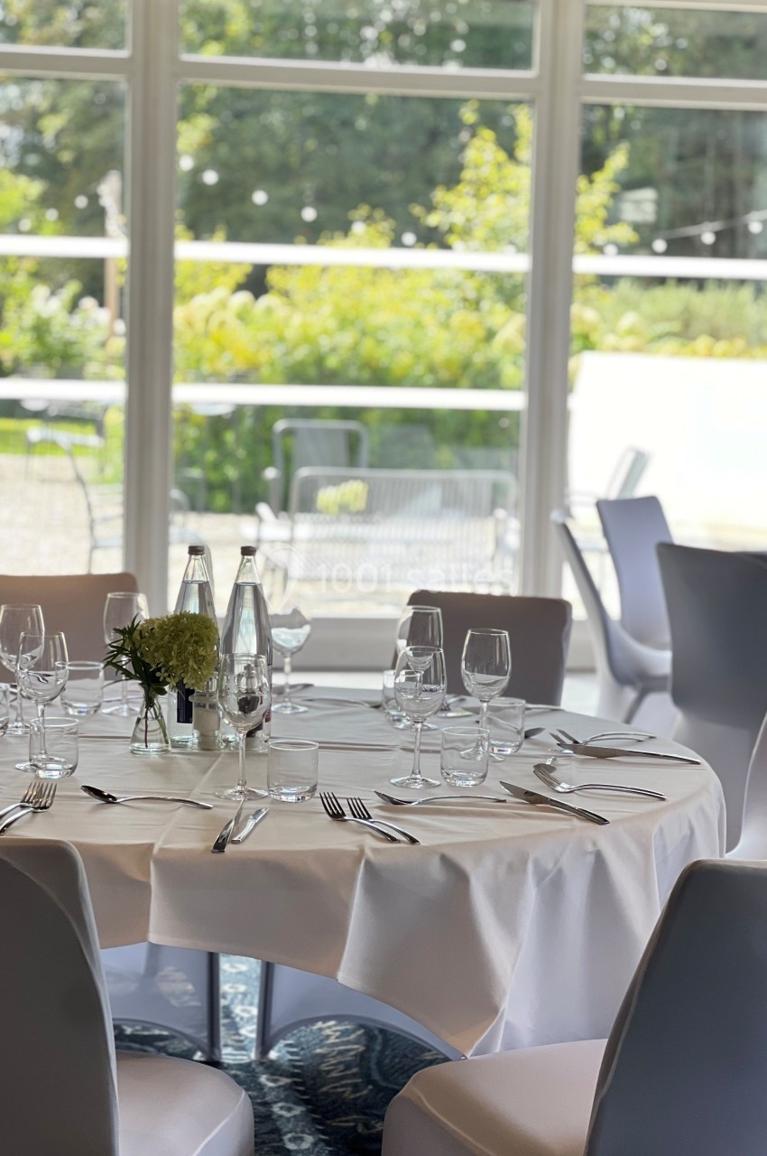 Location salle Marcq-en-Baroeul (Nord) - Mercure Lille Marcq en Baroeul #6 Table dressée avec nappe blanche, verres, couverts et centre de table floral, devant une baie vitrée donnant sur un jardin.
