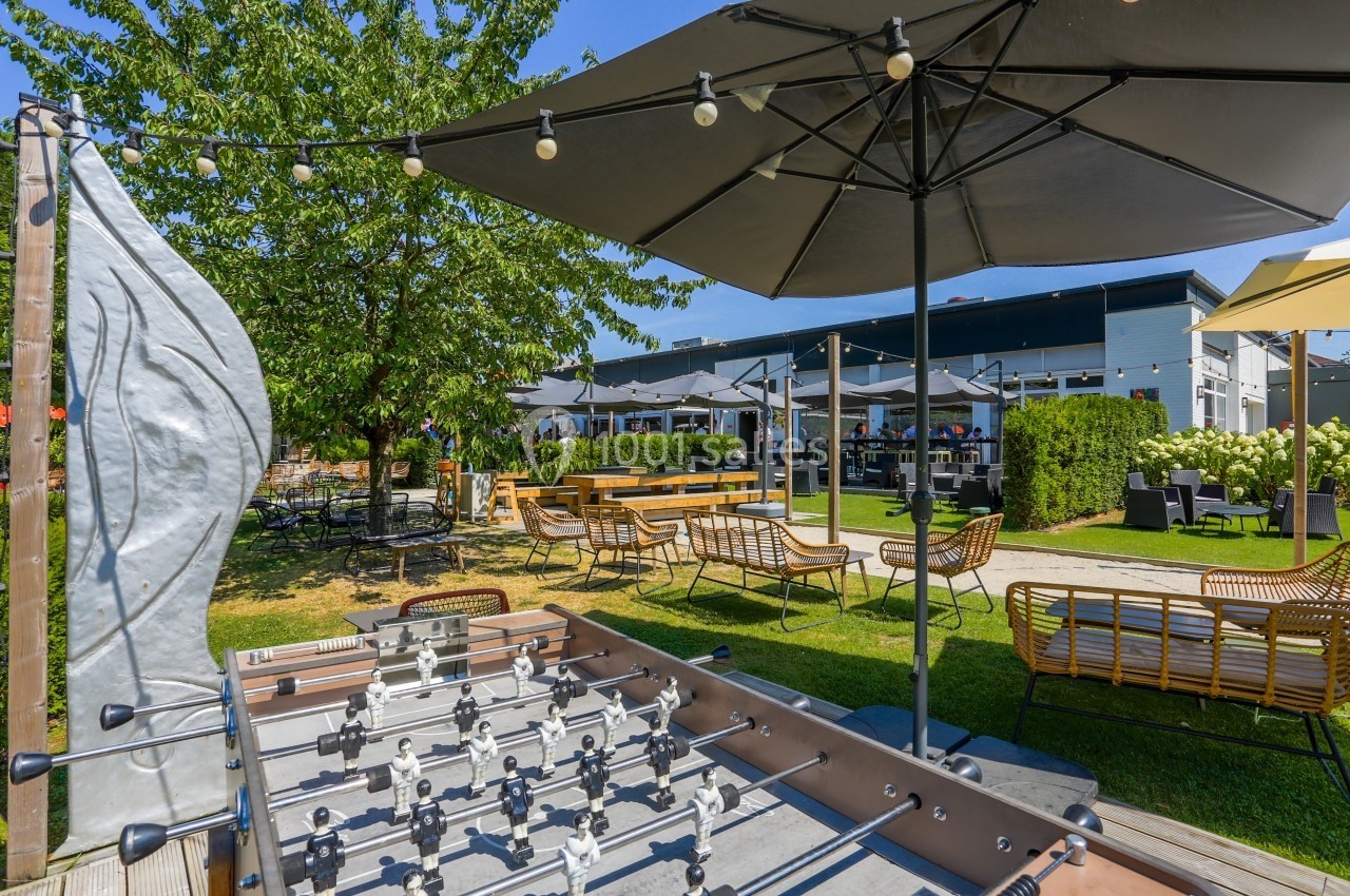 Location salle Marcq-en-Baroeul (Nord) - Mercure Lille Marcq en Baroeul #8 Terrasse extérieure avec baby-foot, parasols, mobilier de jardin et espace verdoyant sous un ciel ensoleillé.