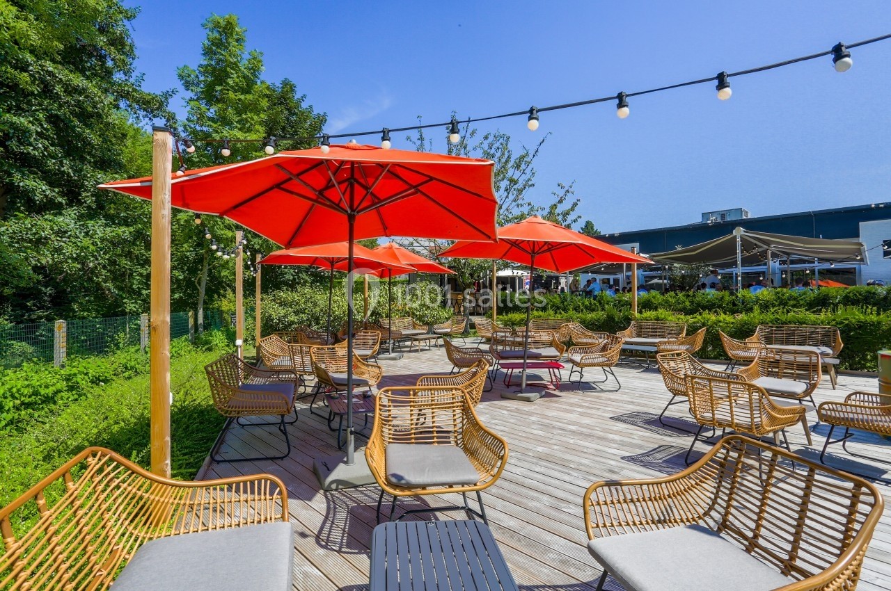 Location salle Marcq-en-Baroeul (Nord) - Mercure Lille Marcq en Baroeul #10 Terrasse en bois avec des fauteuils en rotin, des parasols rouges et des guirlandes lumineuses, entourée de verdure.
