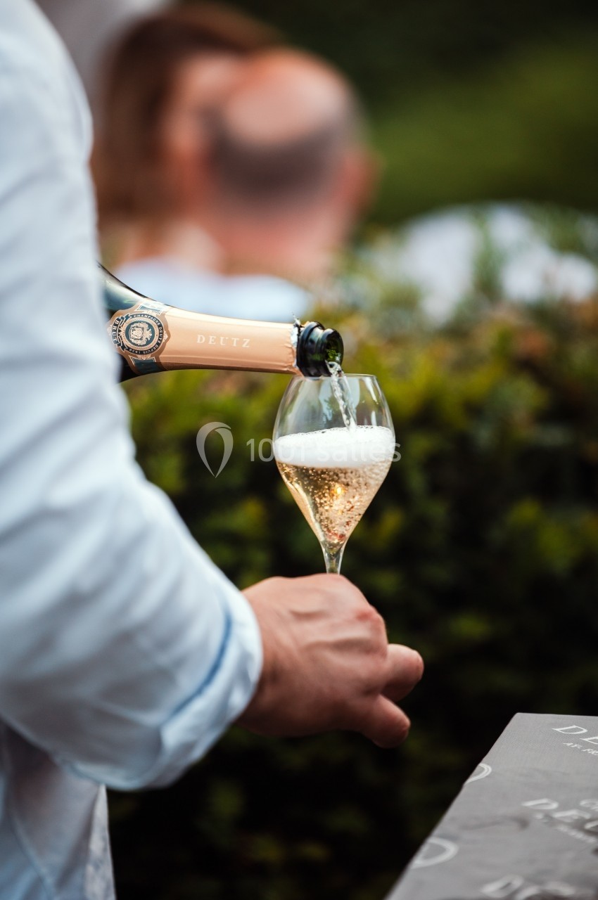 Location salle Marcq-en-Baroeul (Nord) - Mercure Lille Marcq en Baroeul #13 Une main verse du champagne pétillant dans une flûte, avec un arrière-plan flou de verdure et de personnes.