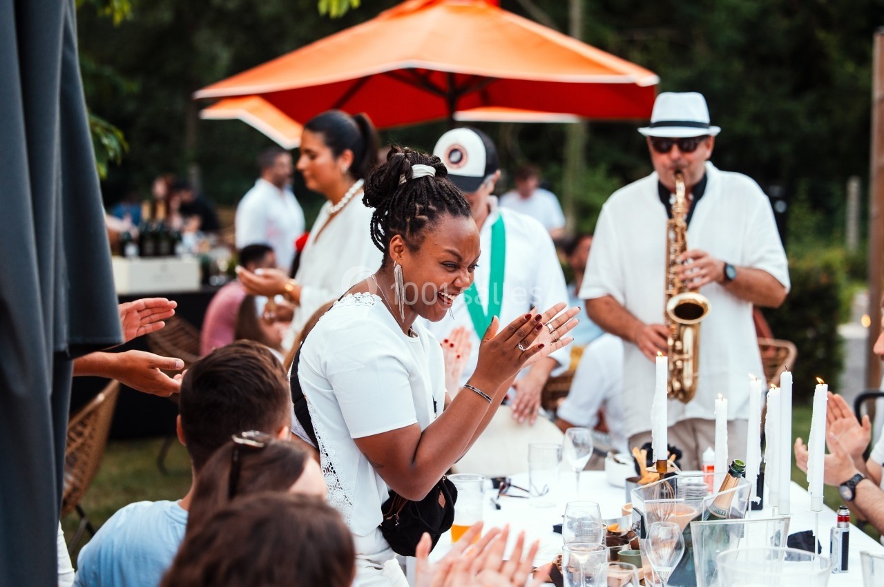Location salle Marcq-en-Baroeul (Nord) - Mercure Lille Marcq en Baroeul #17 Une femme souriante applaudit lors d'un repas en extérieur, avec un musicien jouant du saxophone en arrière-plan.