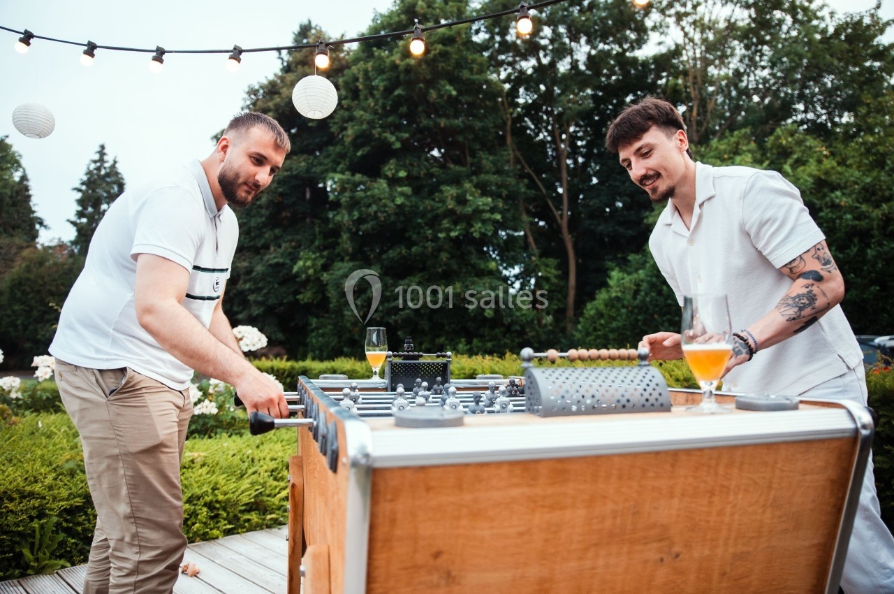 Location salle Marcq-en-Baroeul (Nord) - Mercure Lille Marcq en Baroeul #19 Deux hommes jouent au baby-foot en extérieur, entourés de verdure et de guirlandes lumineuses.