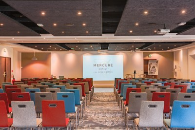 Miniature Location salle Marcq-en-Baroeul (Nord) - Mercure Lille Marcq en Baroeul #23 Salle de conférence avec des rangées de chaises colorées face à un écran de projection allumé.