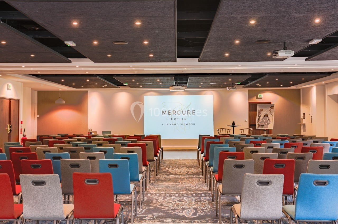 Location salle Marcq-en-Baroeul (Nord) - Mercure Lille Marcq en Baroeul #23 Salle de conférence avec des rangées de chaises colorées face à un écran de projection allumé.