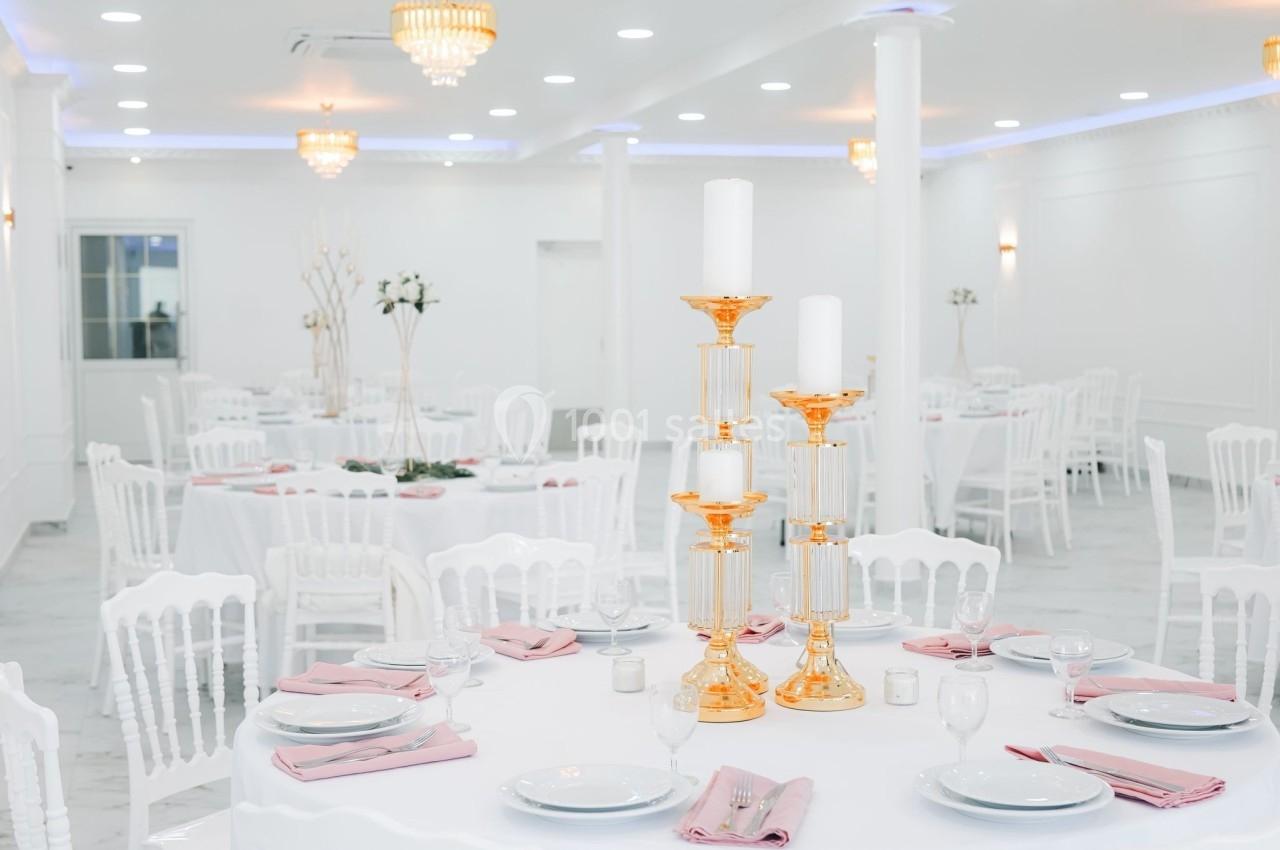Salle de réception lumineuse avec tables rondes dressées, nappes blanches, serviettes roses et chandeliers dorés.