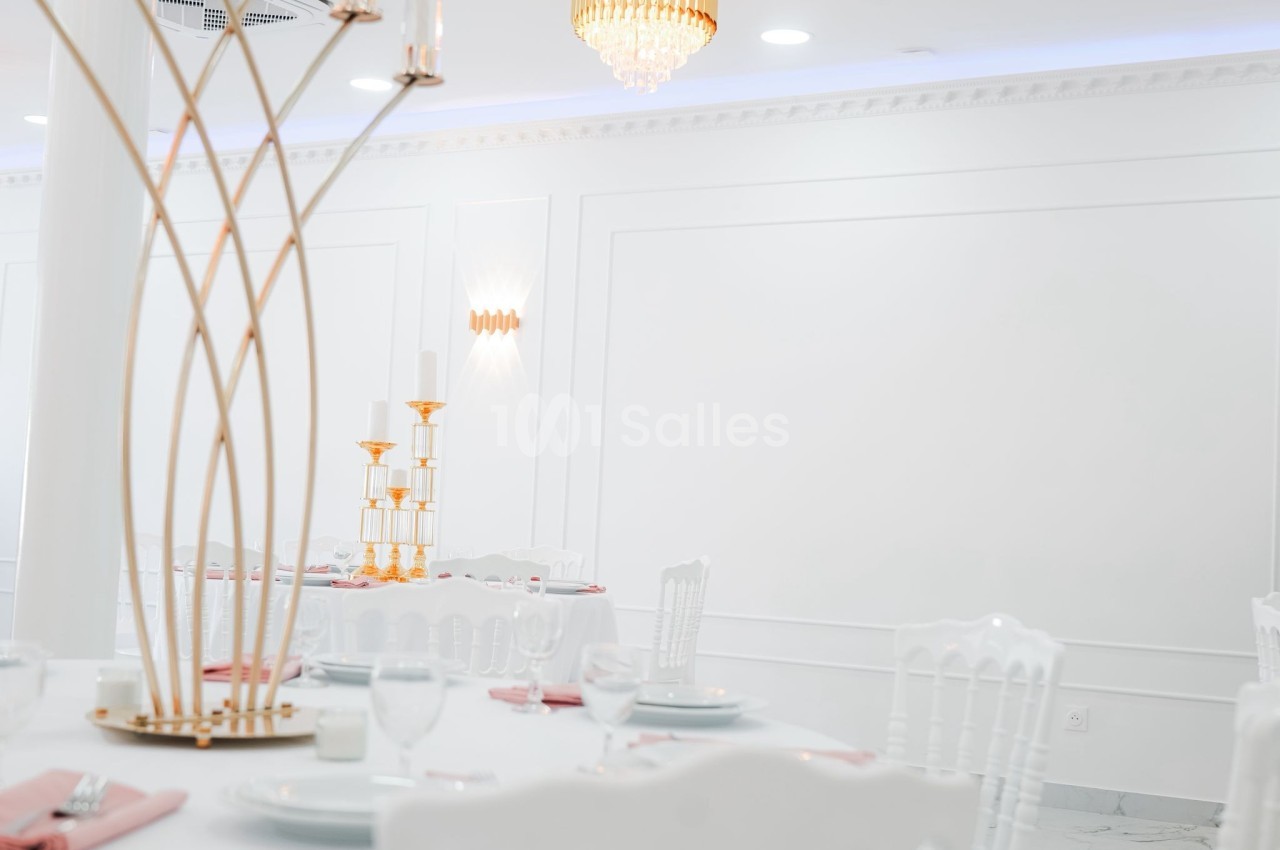 Salle de réception lumineuse avec tables dressées, chaises blanches et décorations dorées élégantes.