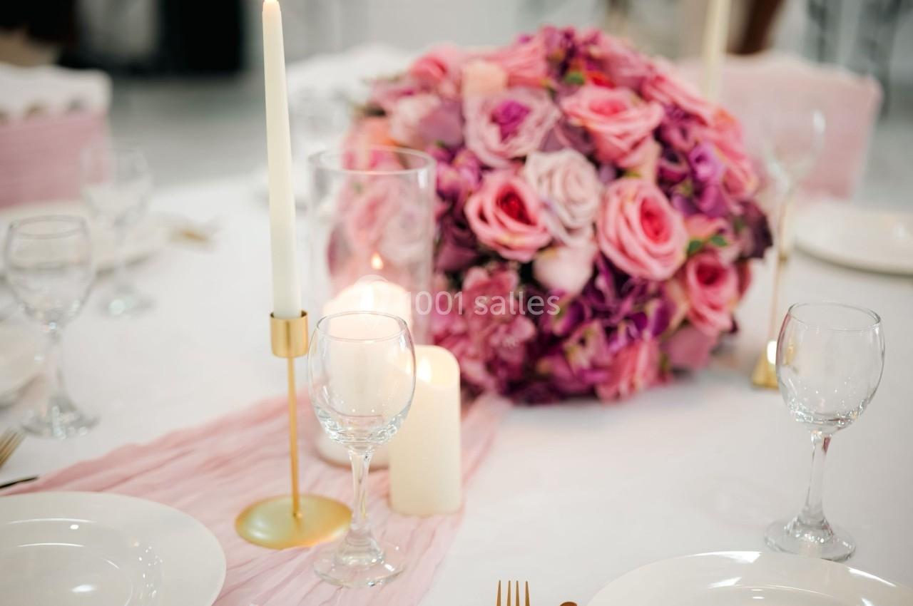 Centre de table avec bouquet de roses roses et violettes, bougies allumées et vaisselle sur une nappe blanche.
