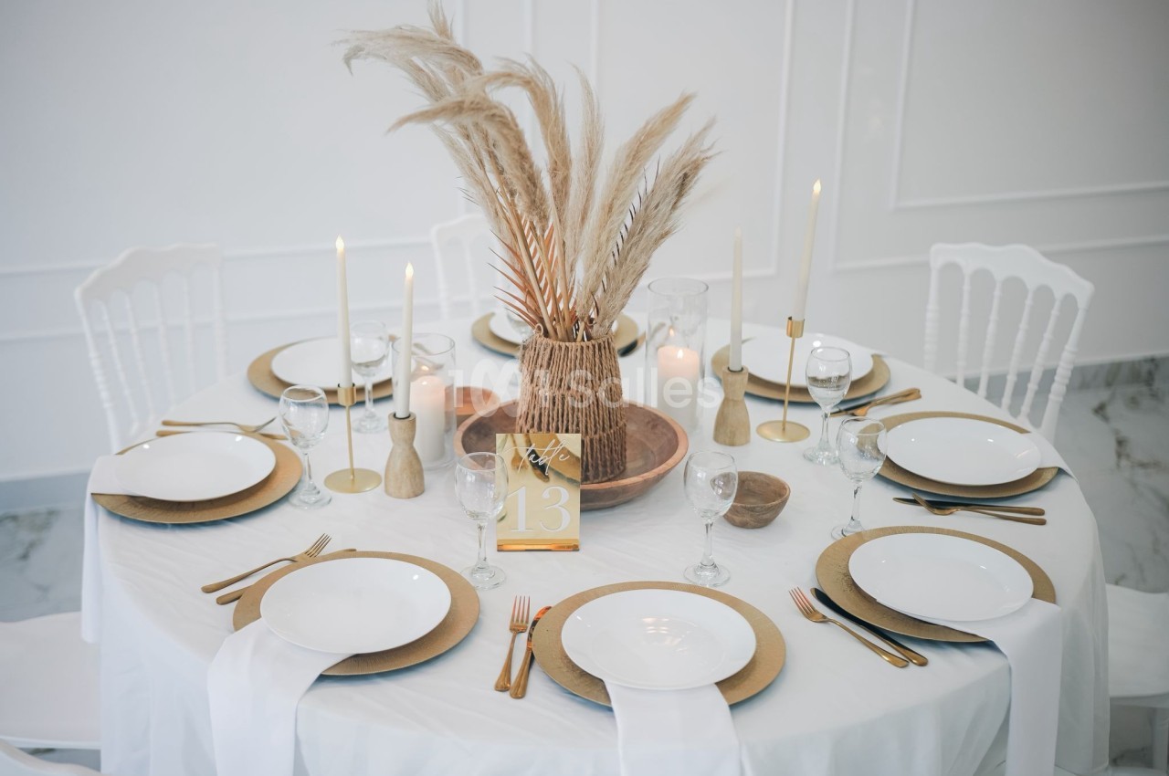 Table ronde élégamment dressée avec nappes blanches, assiettes dorées, bougies et centre de table en pampas.