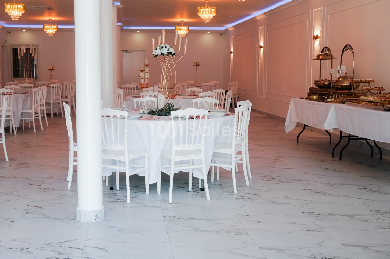 Salle de réception lumineuse avec tables rondes dressées, chaises blanches et buffets alignés sur le côté.