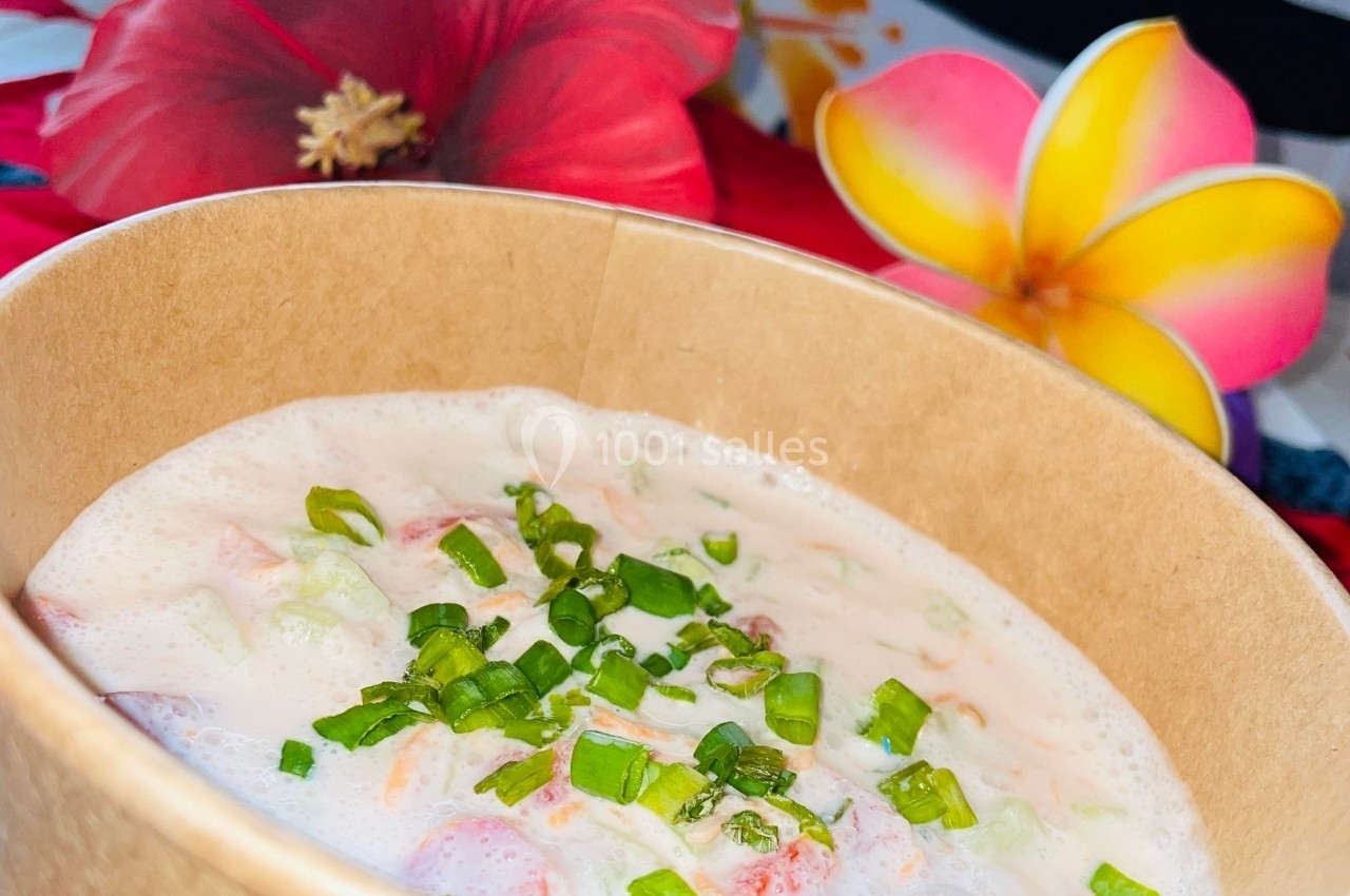 Bol en carton contenant une salade crémeuse garnie de ciboulette, avec des fleurs tropicales en arrière-plan.