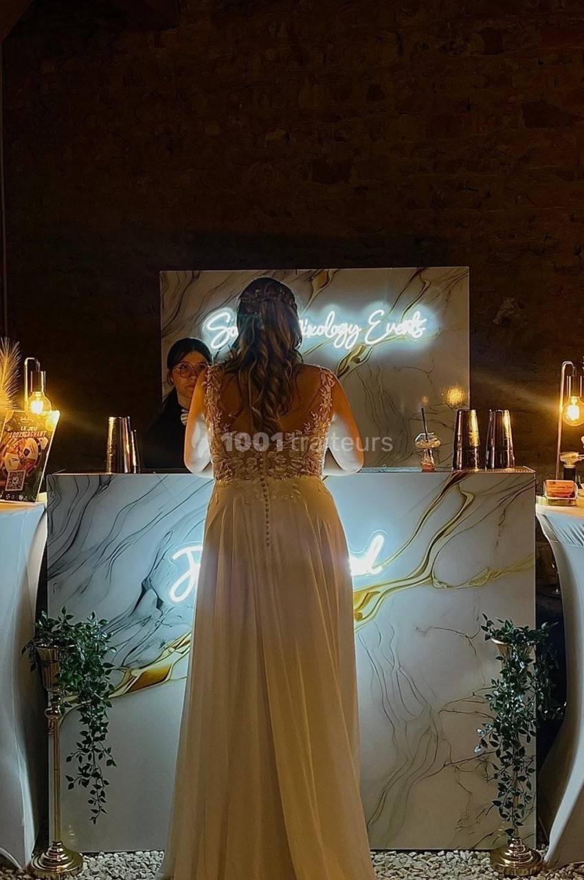 Une femme en robe blanche se tient devant un bar éclairé par des néons et des lampes dans une ambiance tamisée.