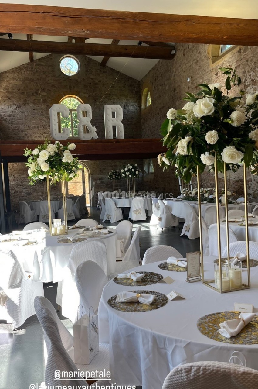 Salle de réception décorée avec des tables rondes, nappes blanches, centres de table fleuris et initiales ’C & R’ en arrière…