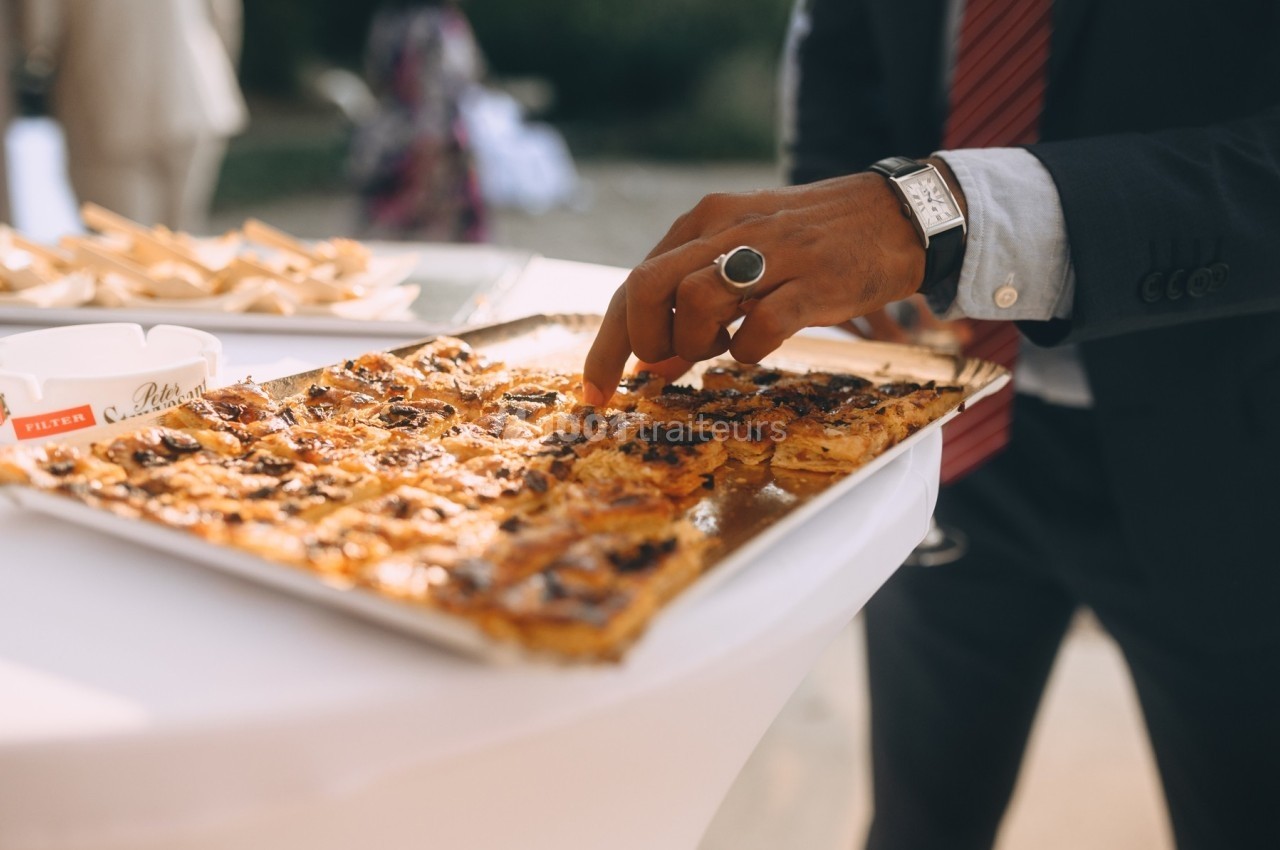 Une personne en costume prend un morceau de tarte salée sur un plateau posé sur une table blanche.
