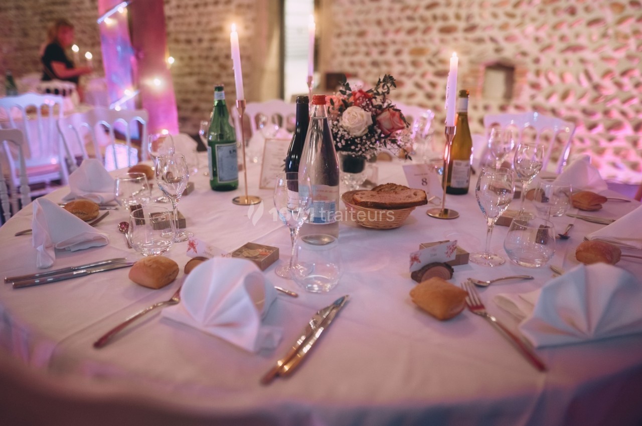 Table de réception dressée avec nappes blanches, couverts, verres, bougies, paniers de pain et décoration florale.