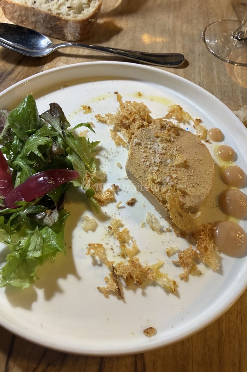 Assiette contenant une tranche de foie gras, salade verte, pickles rouges et points de sauce, sur une table en bois.