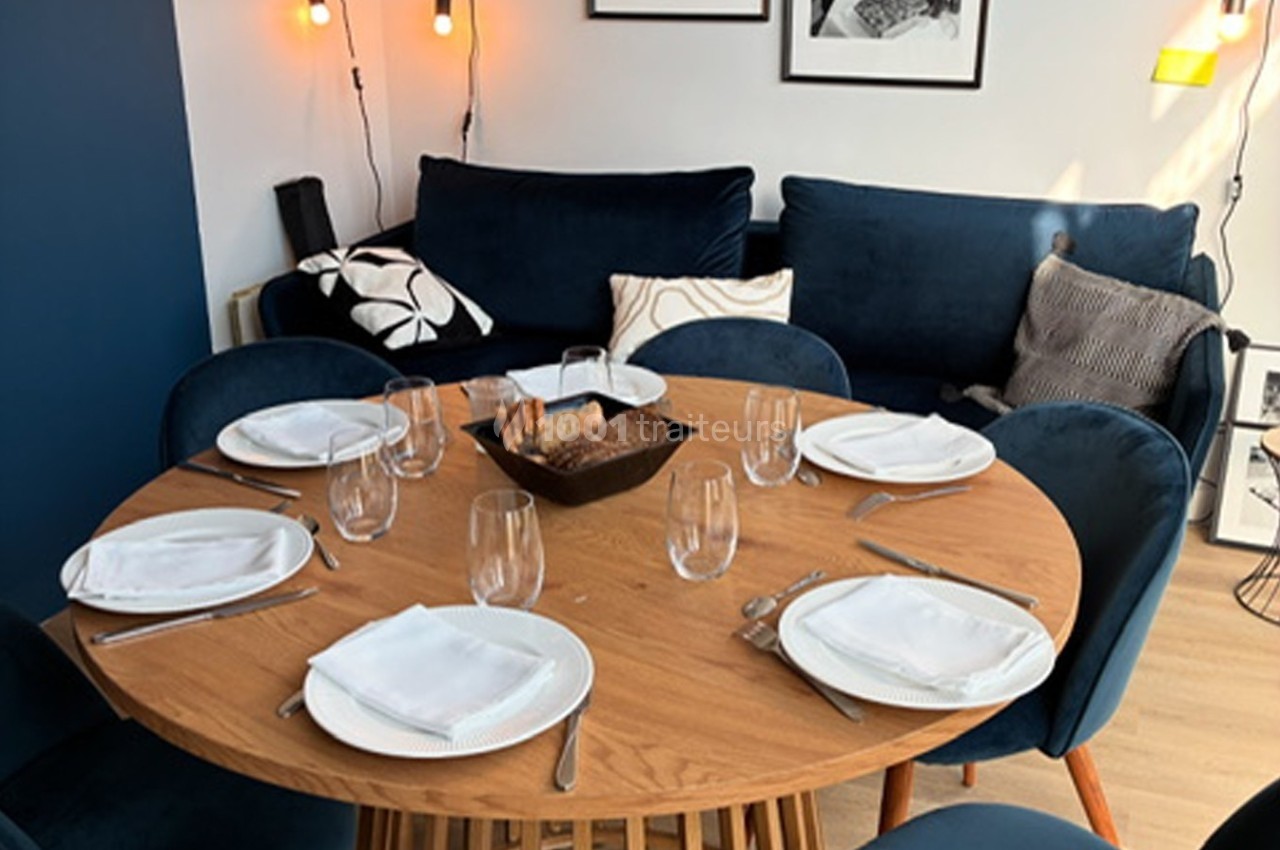 Table ronde en bois dressée pour quatre personnes avec assiettes, verres et serviettes, entourée de fauteuils bleus.