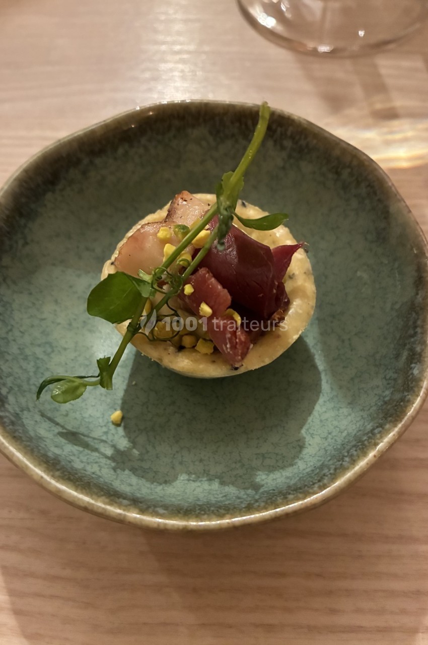 Tartelette garnie de viande séchée, herbes fraîches et éclats de pistache, servie dans une assiette en céramique bleue.