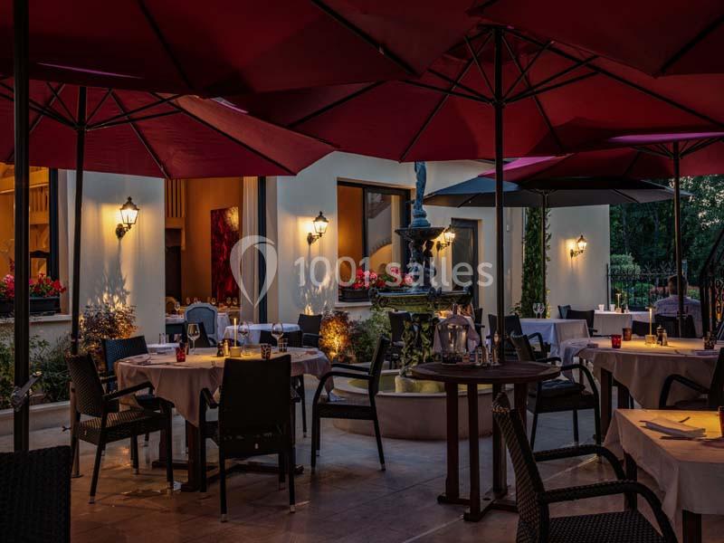 Terrasse d'un restaurant en soirée avec tables dressées, parasols rouges et une fontaine au centre, éclairée par des lampes…