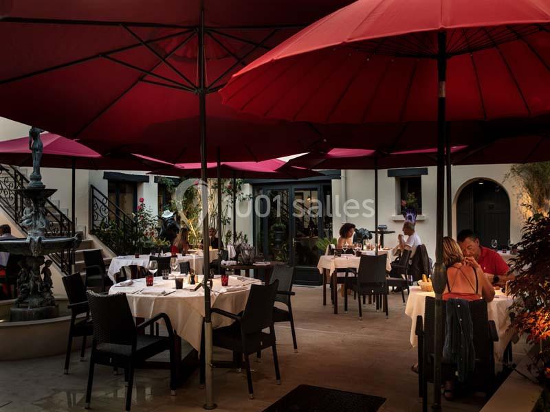 Terrasse d'un restaurant avec des tables dressées, des parasols rouges et une fontaine au centre, ambiance en soirée.