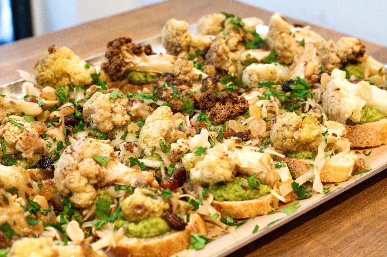 Tranches de pain garnies de chou-fleur rôti, pesto, amandes effilées et herbes fraîches sur une plaque en bois.