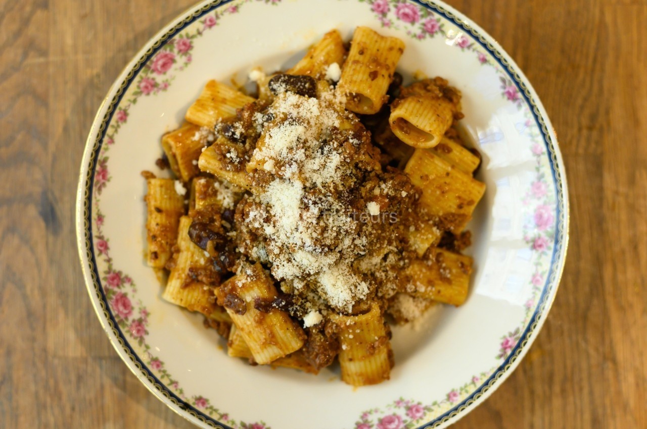 Assiette de rigatoni garnis de sauce tomate à la viande et saupoudrés de fromage râpé, posée sur une table en bois.