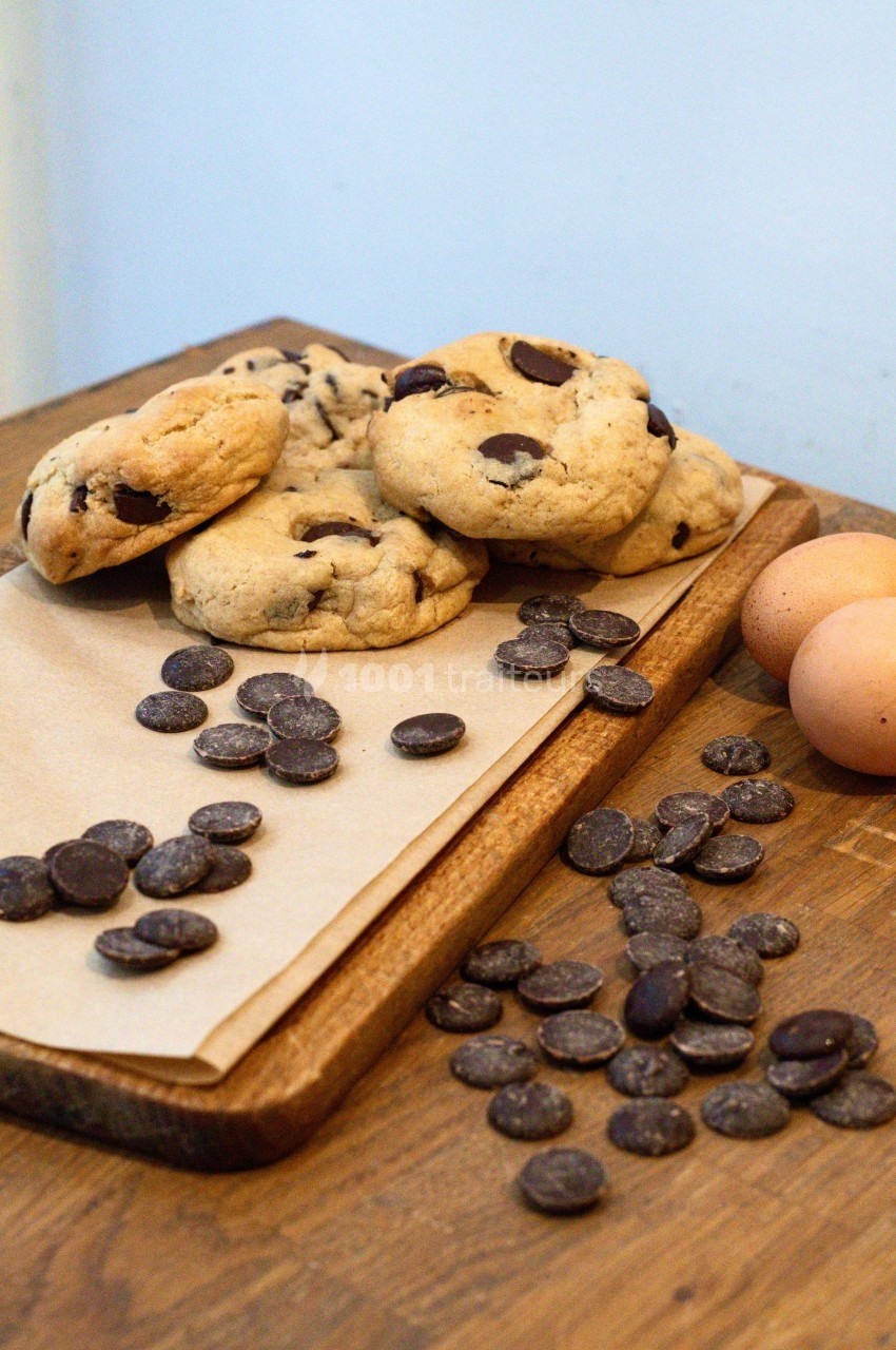 Cookies aux pépites de chocolat sur une planche en bois, entourés de pépites et d'œufs posés à côté.