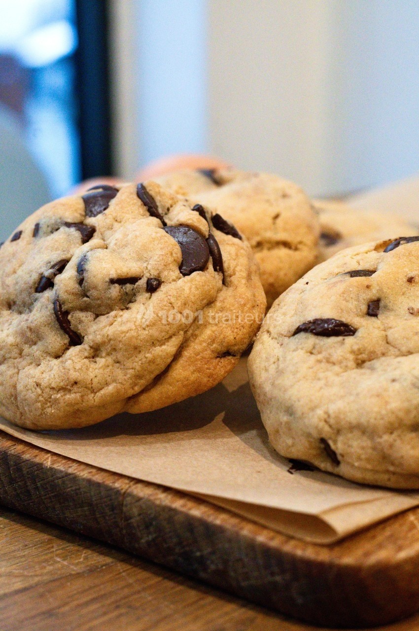 Cookies aux pépites de chocolat posés sur une planche en bois, avec un fond flou d'intérieur lumineux.