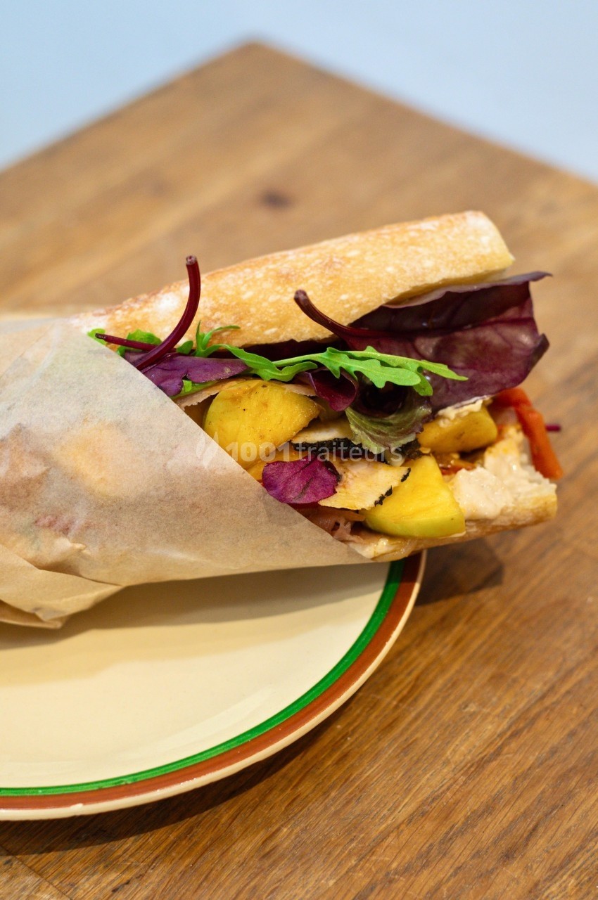 Sandwich garni de poulet, feuilles de salade, morceaux d'ananas et légumes, présenté sur une assiette en bois.