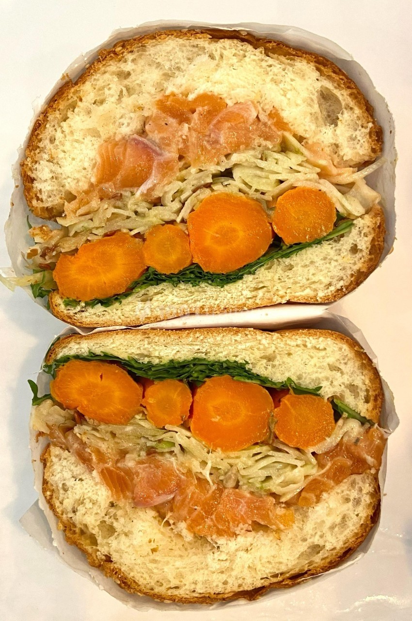 Sandwich garni de saumon fumé, carottes, salade et chou râpé, présenté coupé en deux sur une surface blanche.