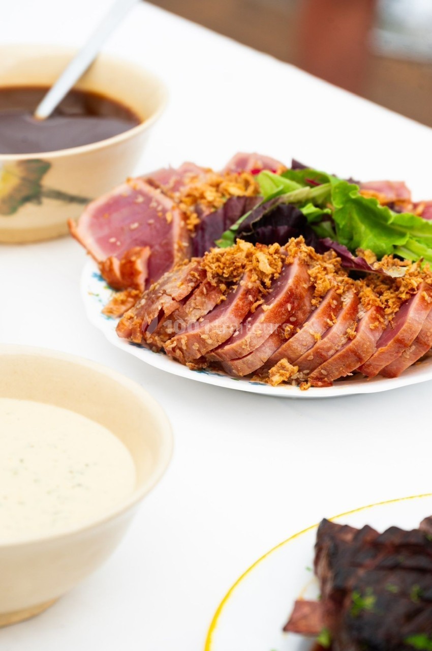 Assiette de tranches de thon mi-cuit garnies de feuilles vertes et d'oignons frits, accompagnée de sauces.