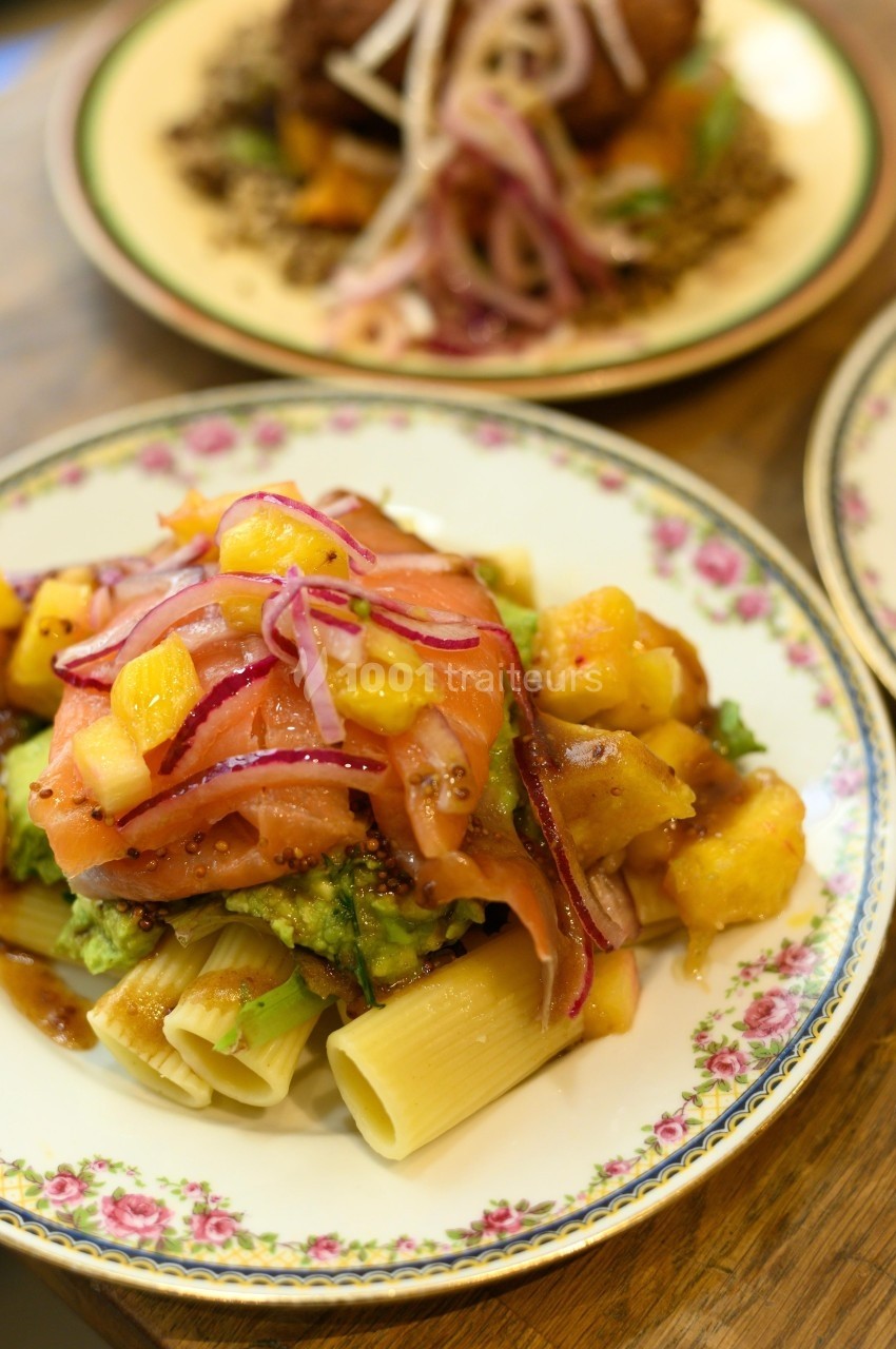 Assiette de pâtes garnie de saumon fumé, mangue, avocat et oignons rouges, servie sur une table en bois.