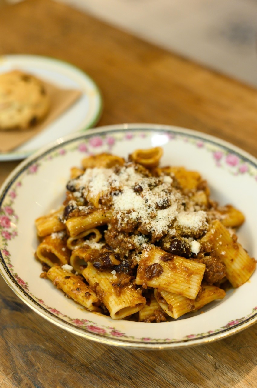 Assiette de pâtes rigatoni à la sauce bolognaise, garnie de fromage râpé, sur une table en bois.