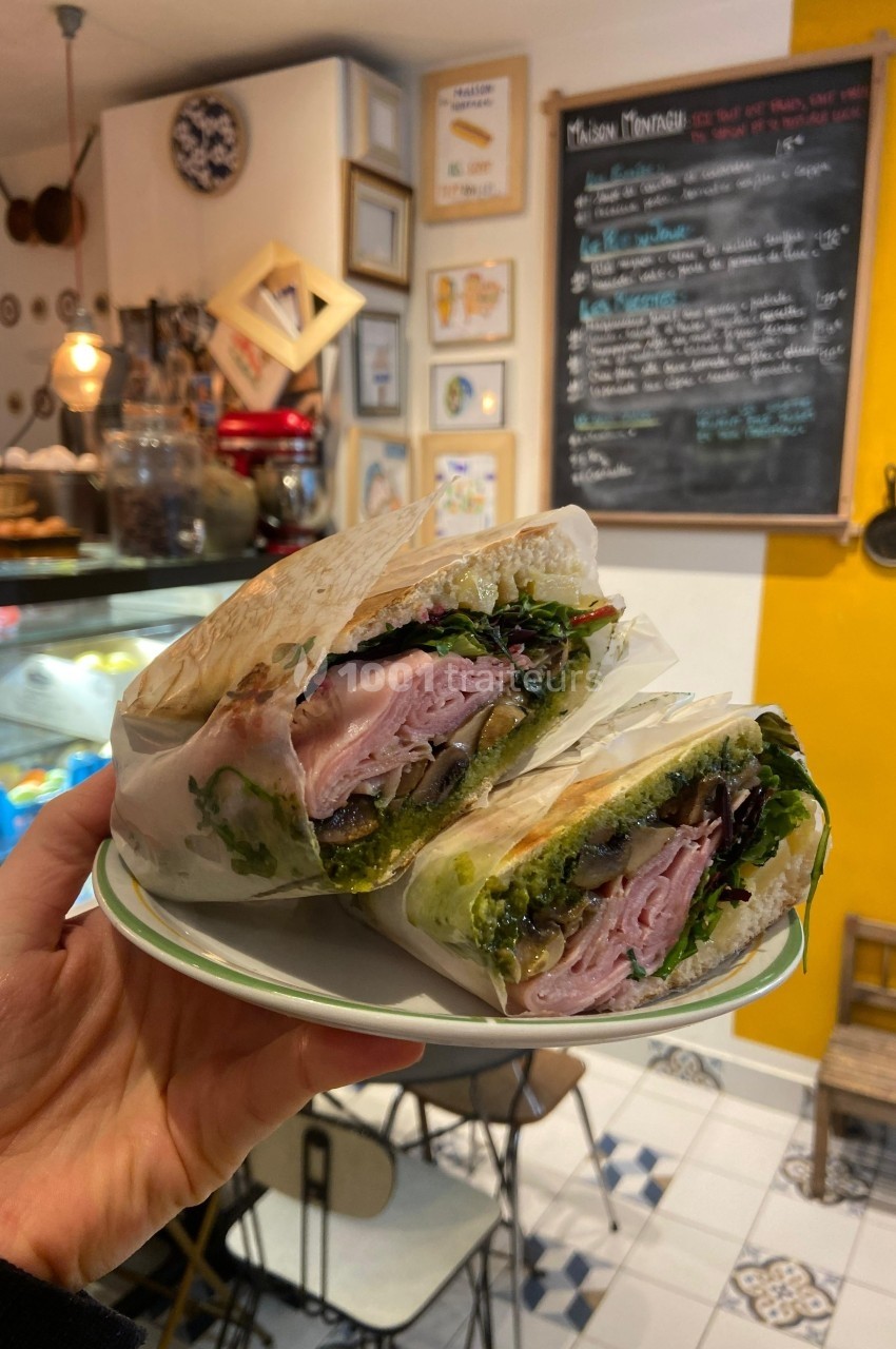 Un sandwich garni de jambon, légumes et sauce pesto, présenté coupé en deux dans un café chaleureux.