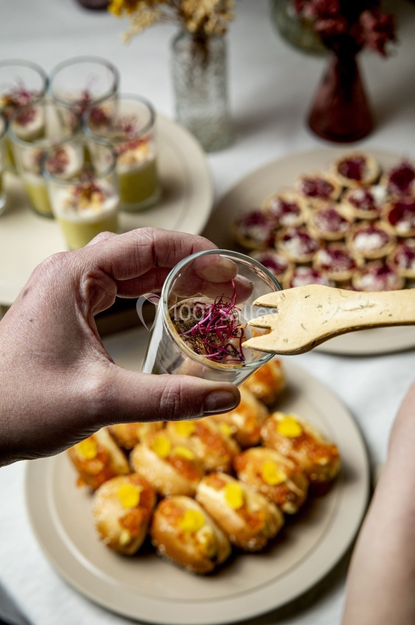 Une main tient un verre de verrine décorée, entourée d'assiettes de bouchées et d'amuse-bouches variés.