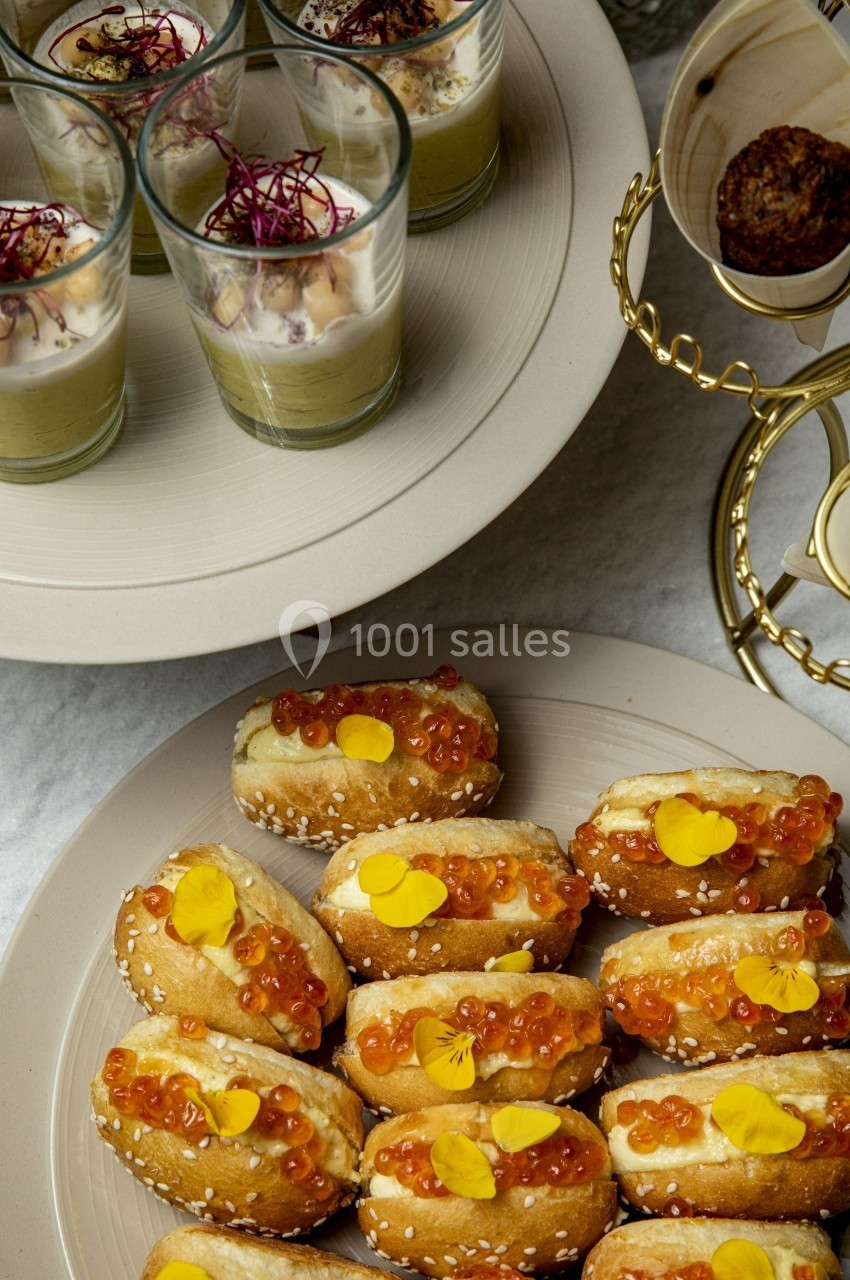 Plateau de mini hot-dogs garnis d'œufs de poisson et fleurs comestibles, avec verrines en arrière-plan.