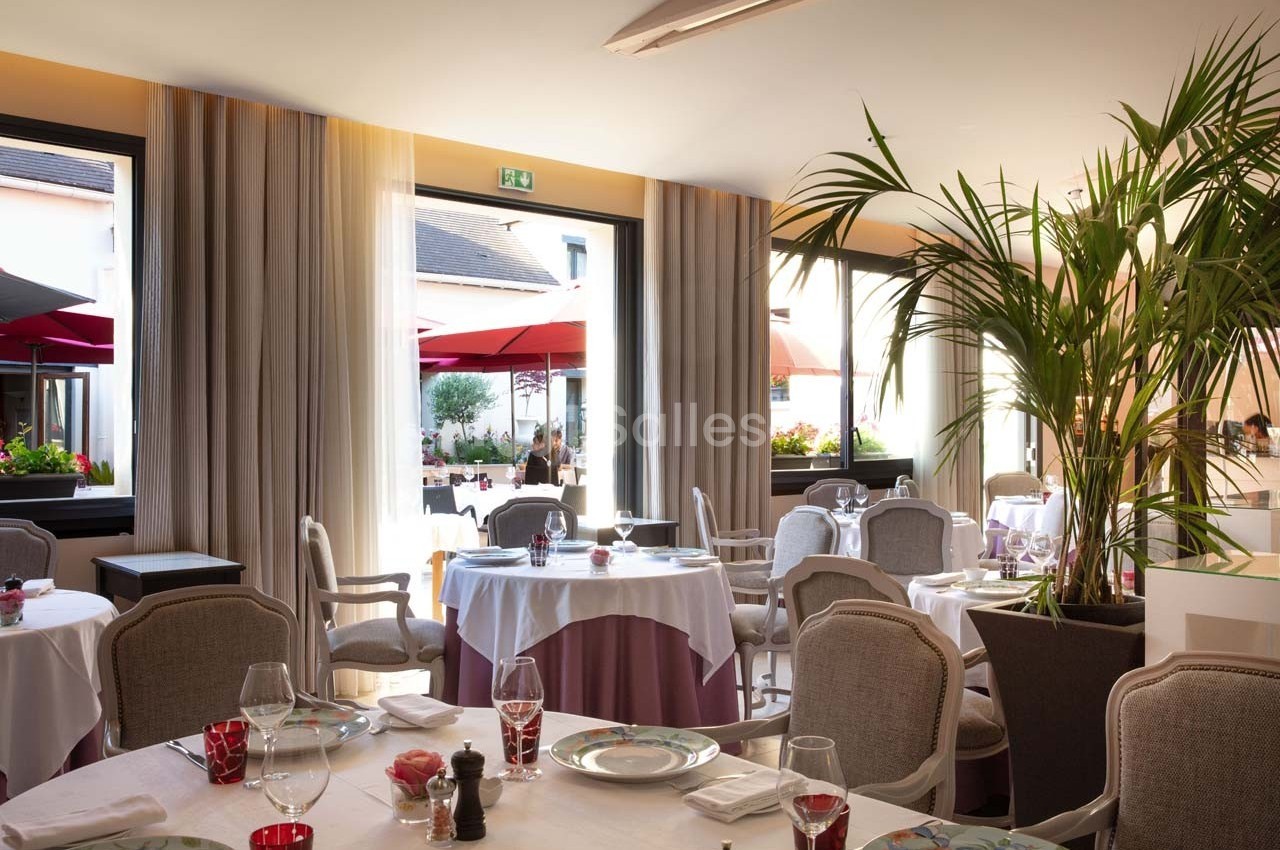 Salle de restaurant lumineuse avec tables dressées, grandes fenêtres ouvertes sur une terrasse avec parasols rouges.
