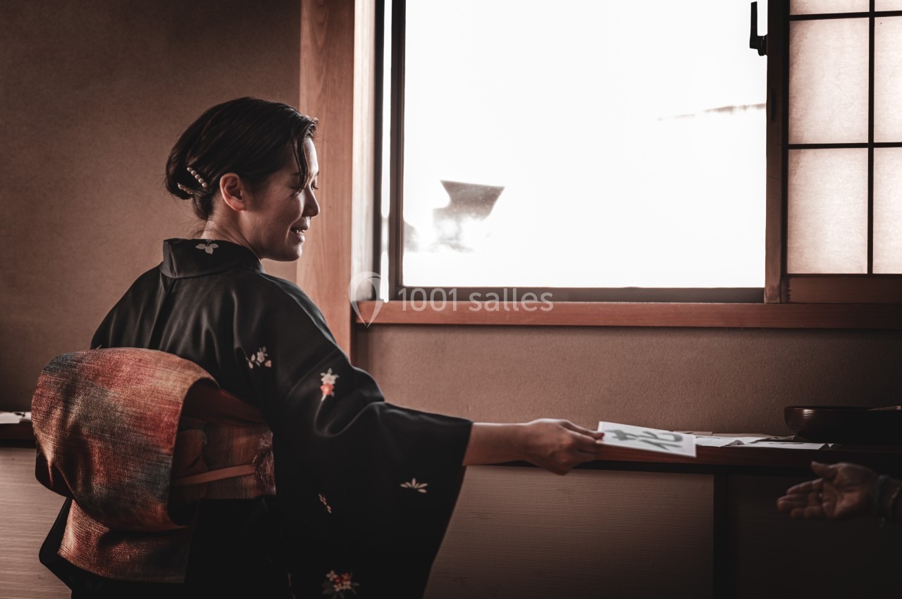 Une femme en kimono tend un document à une autre personne dans une pièce éclairée par une fenêtre.