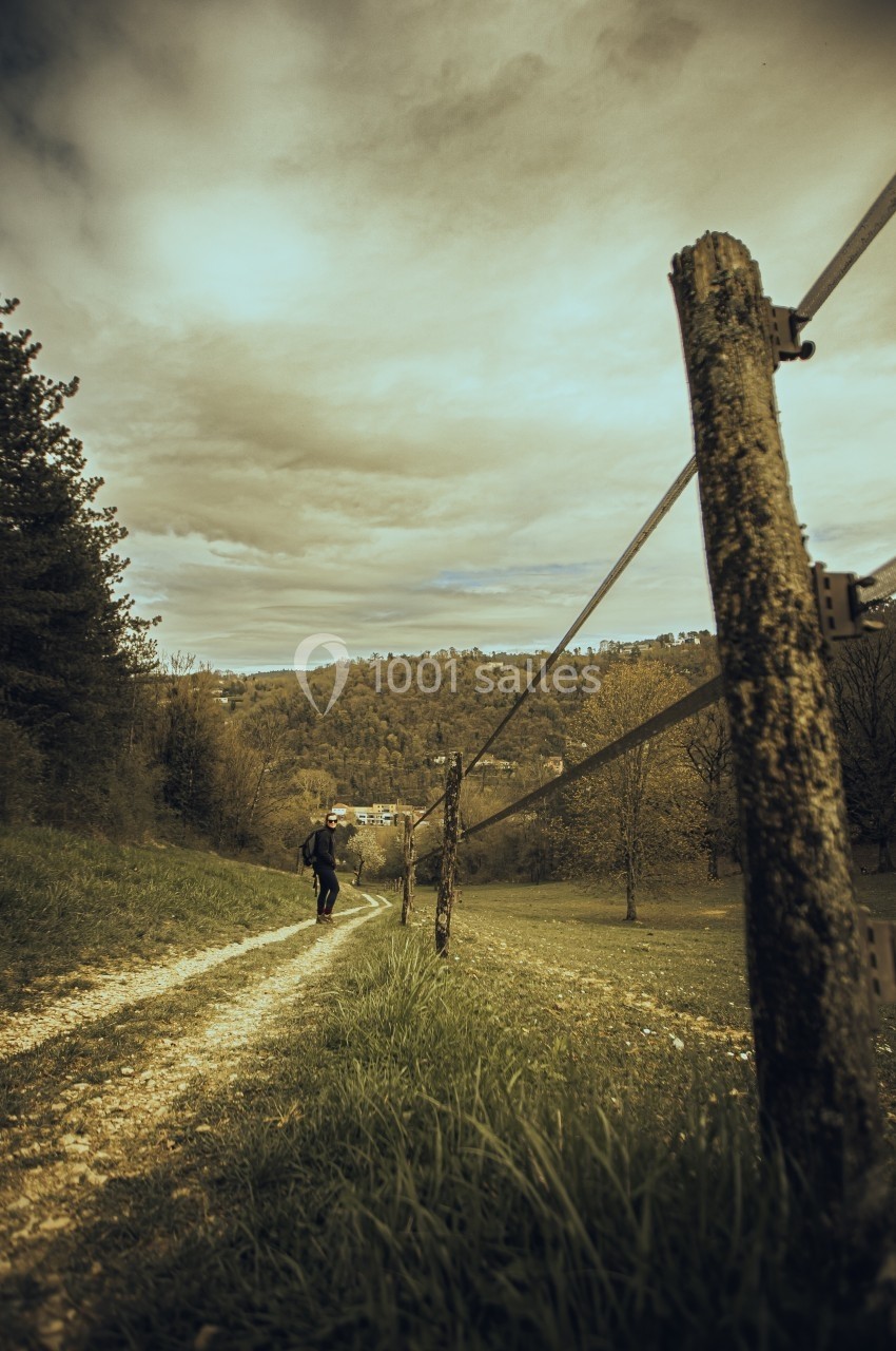 Une personne marche sur un chemin de terre bordé d'herbe et de poteaux, avec une vue sur une vallée boisée.
