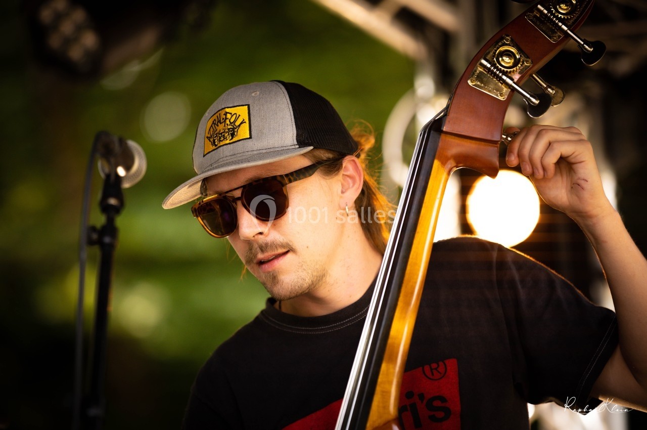 Un musicien joue de la contrebasse sur une scène extérieure, portant des lunettes de soleil et une casquette.