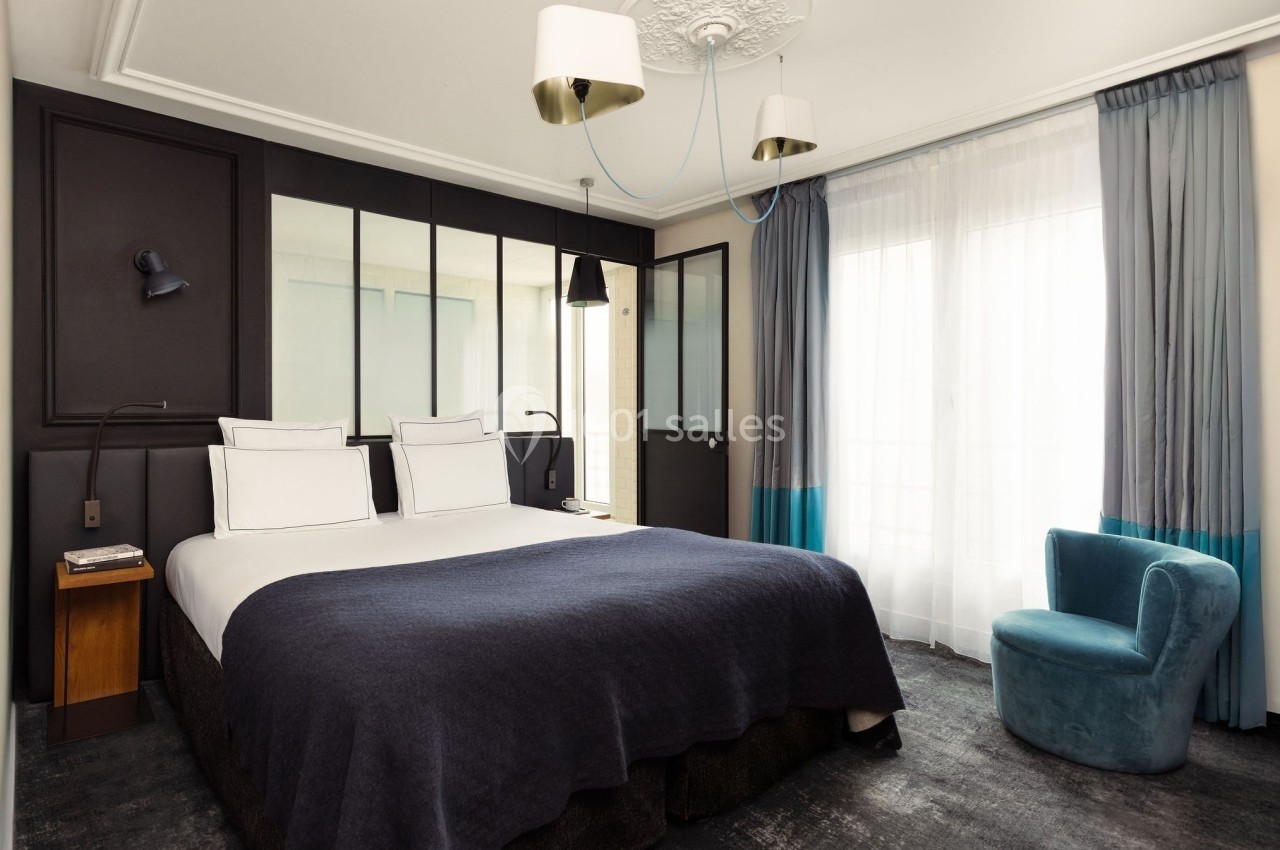 Chambre lumineuse avec un lit double, des murs sombres, une verrière, un fauteuil bleu et des rideaux blancs et bleus.