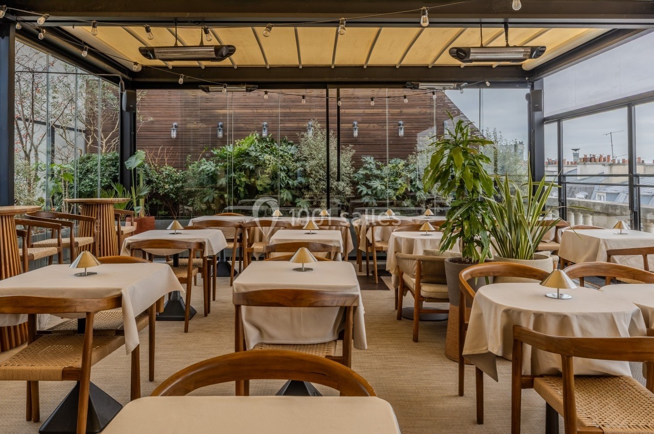 Salle de restaurant lumineuse avec tables dressées, chaises en bois et grandes baies vitrées donnant sur des plantes.