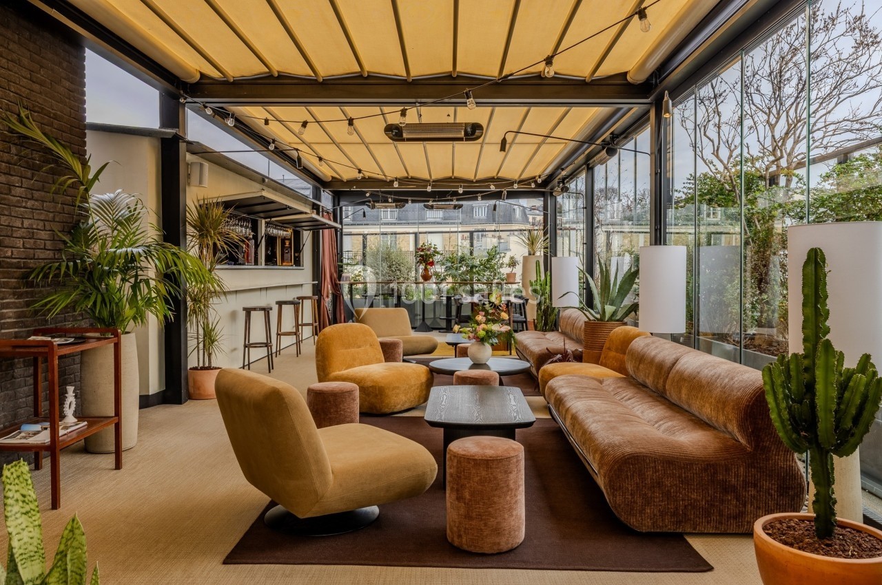 Espace lounge lumineux avec canapés, fauteuils, tables basses et plantes, sous une verrière avec toit ouvrant.