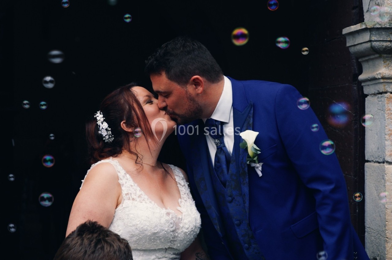 Un couple en tenue de mariage s'embrasse devant une porte, entouré de bulles de savon.